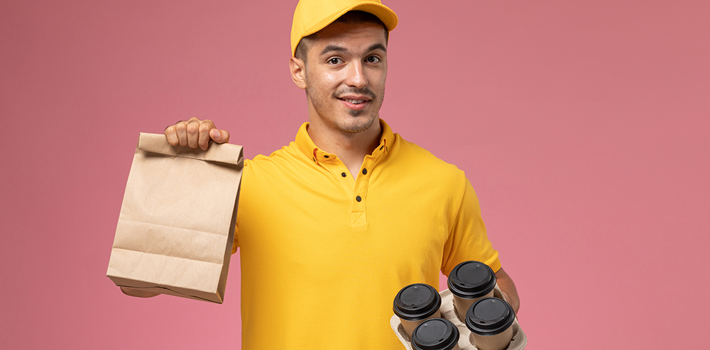 front view male courier in yellow uniform holding food package and delivery coffee cups on the pink background service delivery job uniform