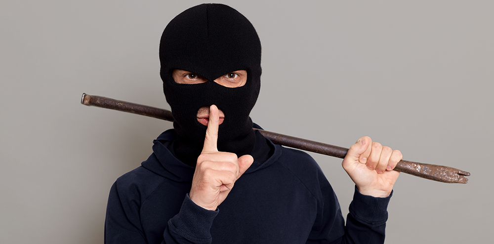 Young guy robber man dressed in a black hoodie with masked face, holds iron crowbar in his hands, shows silence gesture, keeping fore finger near his lips, isolated on gray background