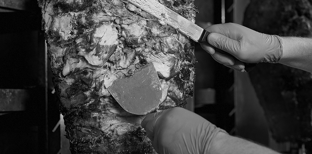 Roasted meat on the bone for the preparation of donors or shawarma. Close-up. The cook cuts the meat fried over charcoal for the preparation of shawarma in pita bread. Fried meat on a skewer for cooking of donors or shawarma. Hands close-up cuts meat in thin slice