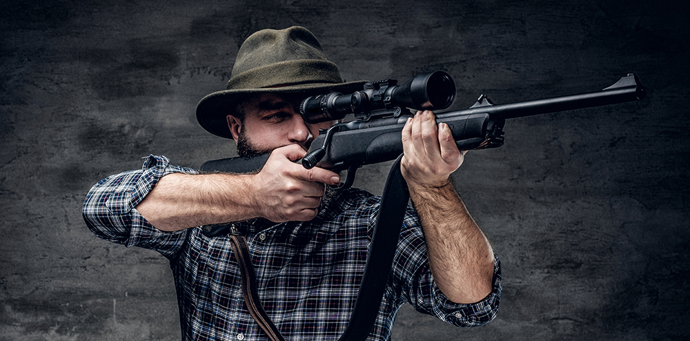 Studio portrait of bearded hunter male wearing a plaid fleece shirt holds a rifle.