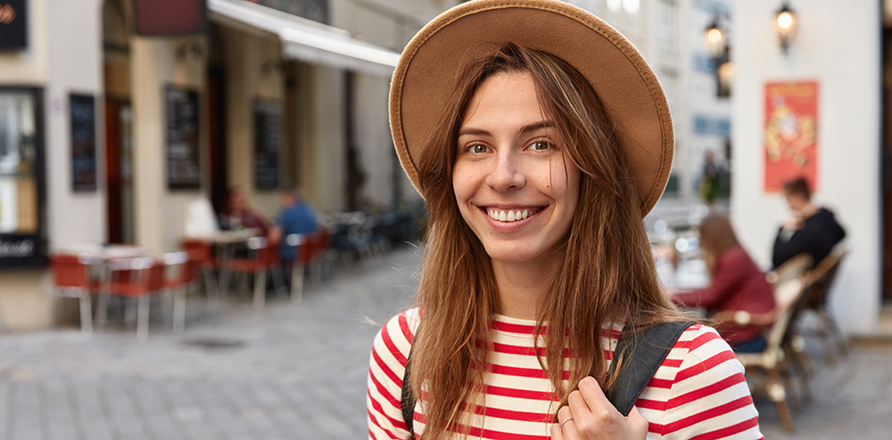 Shot of lovely female smiles gladfully, wears hat and striped jumper, being in good mood as strolls across city, has excursion during trip, poses outdoor, has spare time. People and tourism concept